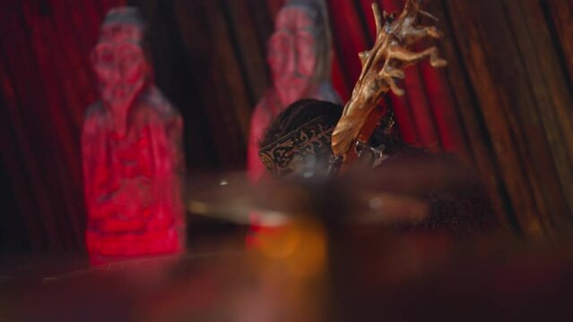 Plump musician plays string instrument with tree root on head in traditional living cabin side view. Authentic native decor of musical instrument