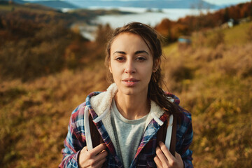 Close up face outdoors portrait young traveler woman with straight look in camera. Tired during mountains hiking, relaxing in nature