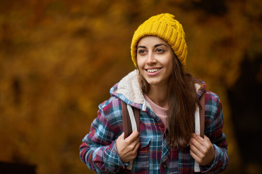 Close up portrait of young happy woman in checkered jacket and yellow beanie with backpack enjoying walking in orange fall forest