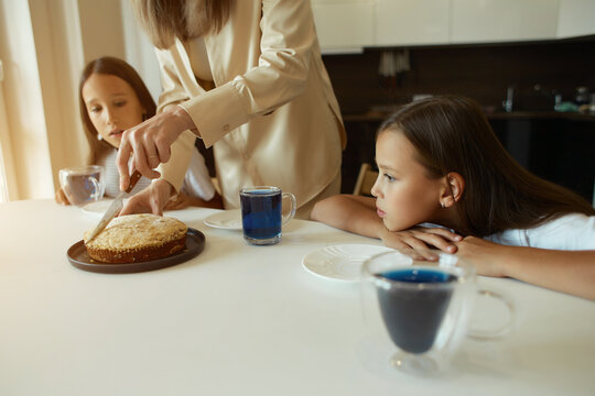 Charming attractive beautiful caucasian kind young smiling mother is drinking tea with her daughters, dressed in casual clothes, sitting in a bright kitchen, cutting pirok