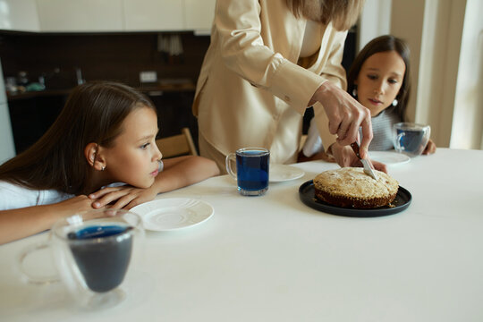 Charming attractive beautiful caucasian kind young smiling mother is drinking tea with her daughters, dressed in casual clothes, sitting in a bright kitchen, cutting pirok