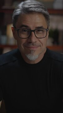 Confident older white man at home, sitting on couch in dark living room. Portrait of happy, mature age, middle age, mid adult caucasian man in 50s, smiling, wearing glasses.