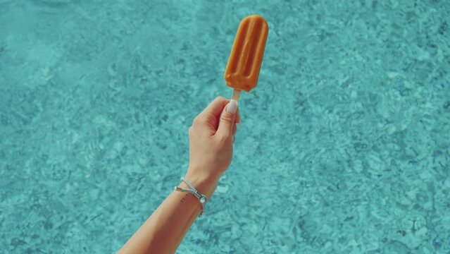 Woman hand holding orange ice cream with a swimming pool in the background. Summer days, parties, enjoying vacation. 