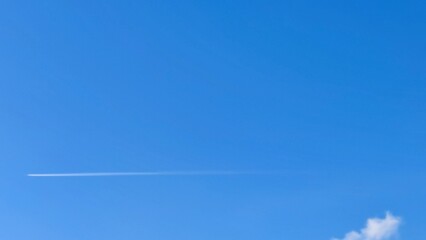 Light blue sky and airplane. In a clear blue sky with sparse clouds in the distance, an airplane flies, leaving a white long straight trail behind it.