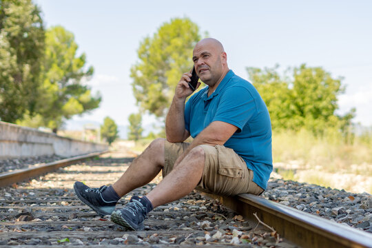 Overweight Middle-aged Man Sitting On The Train Track Talking On The Phone.