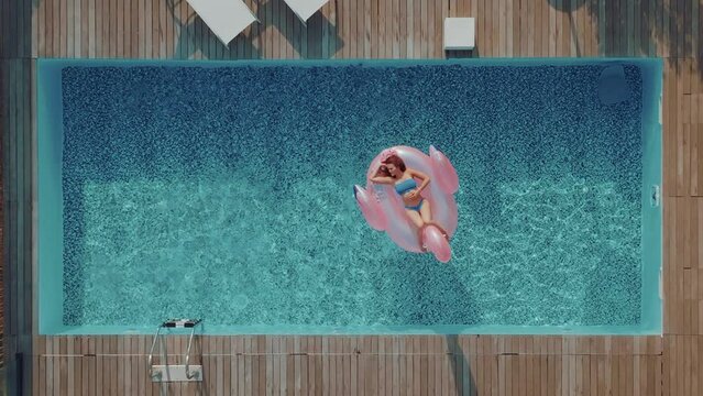 Young tourist woman with long hair is sunbathing on an inflatable pink flamingo in the middle of a swimming pool on a hot summer day. Female relaxing by the pool on a sunny day. Smiling on vacation.