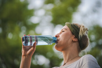 closeup o f a young healthy alternative & spiritual looking woman drinking fresh detox water from a blue plastic bottle outside in the park with a green refreshed nature forest background