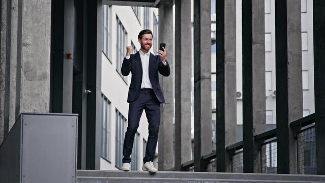 Young stylish businessman stands against backdrop of office building reads good news on telephone celebrates victory rejoices in success business promotion achievement excellent result receives reward