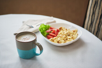 Cappuccino coffee in cup and plate with scrambled eggs and tomatoes. Breakfast on the table