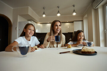 Charming attractive beautiful caucasian kind young smiling mother is drinking tea with her daughters, dressed in casual clothes, sitting in a bright kitchen, cutting pirok
