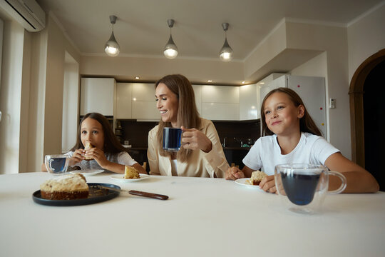 Charming attractive beautiful caucasian kind young smiling mother is drinking tea with her daughters, dressed in casual clothes, sitting in a bright kitchen, cutting pirok