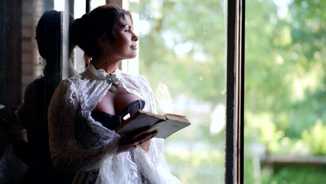 sexy woman in vintage dress with open decollete is reading book and looking at open window of village house
