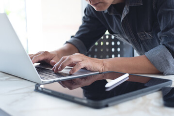 Close up of asian casual business man surfing the internet, online working on laptop computer with digital tablet on desk at home office, paperless office, telecommuting, working from home