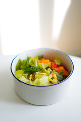 Bowl of vegan salad with tomato, lettuce, basil and mustard with oil and salt on light table with flower