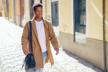 Young black man walking down the street carrying a briefcase and a smartphone.