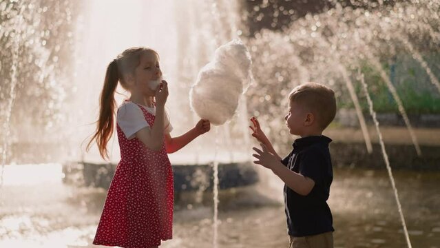 Little girl with brother eats cotton candy near fountain
