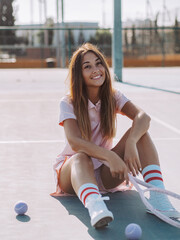 Hispanic female smiling, sitting on a tennis court, holding a tennis racket
