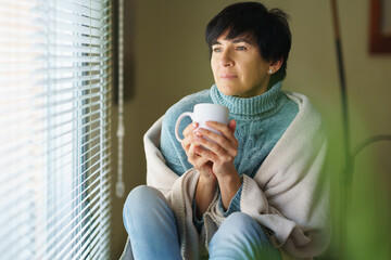 Mid adult woman drinking coffee and looking out of the window in winter day.