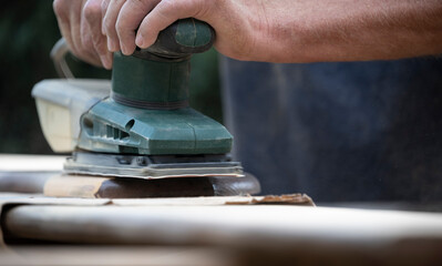 Close-up of an amateur craftsman with an orbital sander in his hand touching up a wooden board in the garden