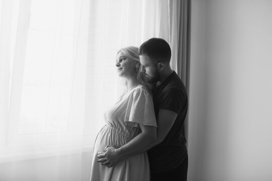 A man hugs his pregnant wife from behind and touches his belly near the window of the house. Pregnant couple. Black and white photo.