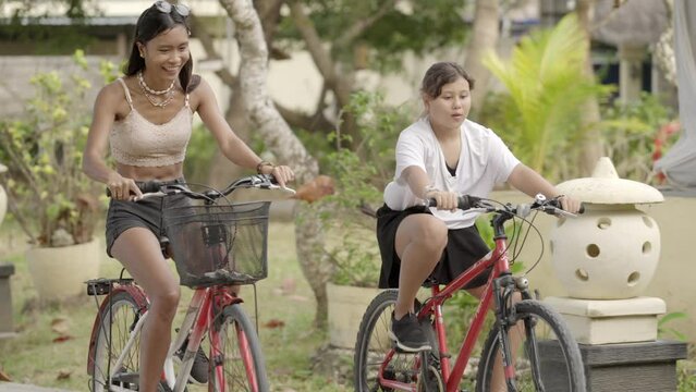 two young asian girls riding bicycle while talking and laughing on a sunny day in slow motion