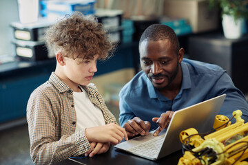 Portrait of black male teacher helping young boy building robot during engineering class in school...