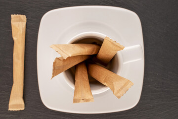Several sticks of sugar in paper bags with a white saucer and a cup on a slate stone, close-up, top...
