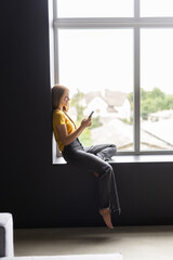 Attractive young woman sitting on a windowsill at home and texting on her phone