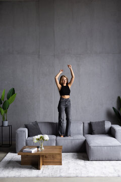 Young Woman Dancing On Yellow Couch In Living Room Near Coffee Table With Laptop