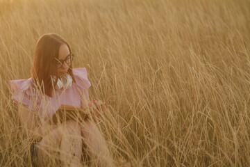 Thoughtful young female in casual clothes and eyeglasses sitting in dry grass and reading a book at sunset