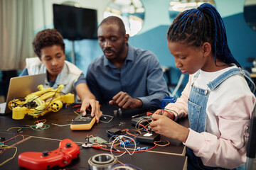 Side view portrait of young black girl building robots with male teacher in background during...