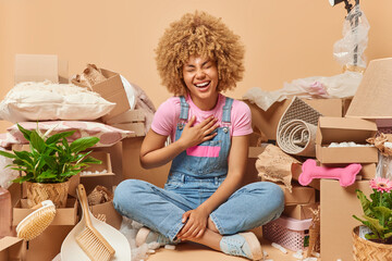 Overjoyed curly haired woman sits crossed legs keeps hand on chest poses on floor in messy room surrounded by cardboard boxes full of household items isolated over beige background. Moving day