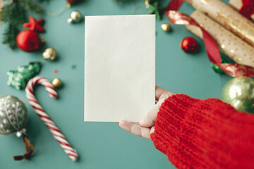 Hand holding christmas card mock up. Empty greeting card in woman hand on background of stylish christmas decorations on green background. Postcard template, space for text. Happy Holidays!
