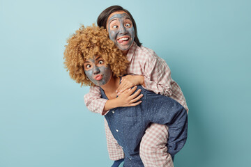 Indoor shot of carefree positive mixed race young women make funny grimaces apply beauty mask take care of skin dressed in slumber suits ride on back of each other isolated over blue background