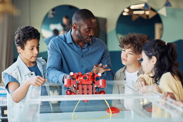 Portrait of black male teacher demonstrating robotic boat to diverse group of children in...