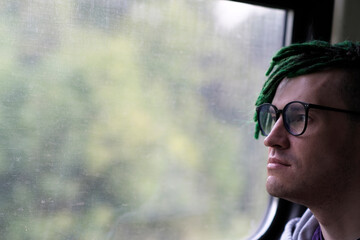 Portrait of young man with green dreadlocks in glasses riding on train, looking at nature. Close up.