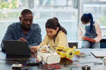 Portrait of Asian teenage girl building robots in engineering class and using laptop with male...