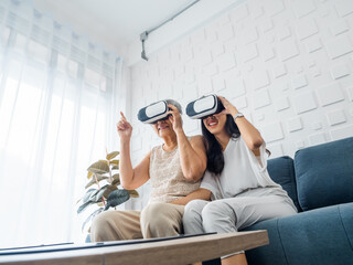 Asian young woman, happy daughter and her mother, old senior female wearing VR glasses, enjoy 3d game online together in white room. Mom and daughter with reality virtual technology lifestyle concept.