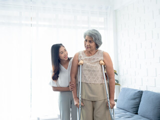 Elderly woman trying to walk on crutches standing held and supported in arms by young Asian female carefully in recovery room, helping old women, health care, senior therapy patient at home concept.