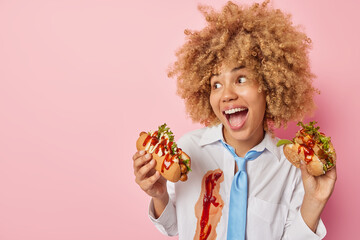 Cheerful woman exclaims loudly looks happily eats fast food holds hot dog and burger wears formal shirt and tie isolated over pink background blank space for your advertising content. Binge eating