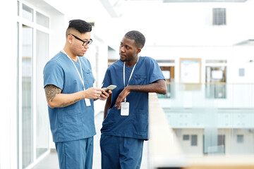 Portrait of two multiethnic doctors wearing uniform standing in hall of modern clinic nd chatting,...