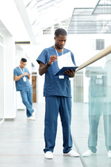 Vertical full length portrait of young African American doctor holding clipboard while standing in hall of modern clinic