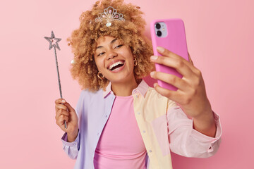 Lovely positive curly haired young woman smiles broadly clicks selfie via smartphone makes photo...