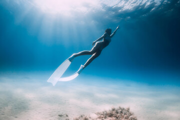 Attractive lady freediver underwater glides with white fins in transparent ocean with sun rays