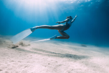 Freediver underwater glides with white fins over sandy bottom. Attractive woman freediver in blue ocean with sun rays