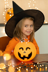 Little girl in black witch hat looks at camera and smiles. Happy child celebrats Halloween with lantern pumpkin