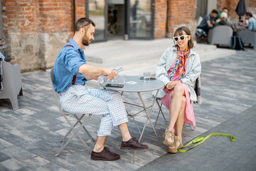 Fototapeta premium Stylish couple of colleagues have a conversation while sitting at cafe terrace in modern office district. Couple spending time together drinking water by the table outdoors