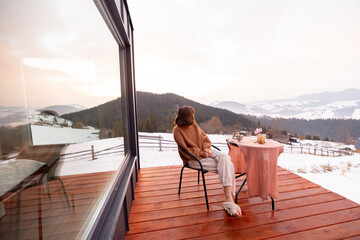 Woman having a breakfast while resting on terrace of tiny house in the mountains, enjoying beautiful landscape during sunrise. Concept of small modern cabins for rest and escape to nature