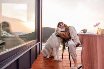 Woman hugs her dog while resting on terrace of tiny house in the mountains, enjoying beautiful landscape during sunrise. Concept of small modern cabins for rest and escape to nature