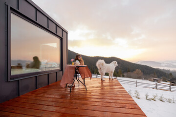 Woman with her dog resting on terrace of tiny house in the mountains, enjoying beautiful landscape during sunrise. Concept of small modern cabins for rest and escape to nature